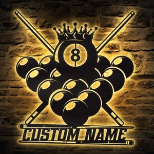 May include: Black metal billiards sign with a crown on the 8-ball, crossed cues, and a rack of billiard balls. The sign has the text "CUSTOM NAME" and is illuminated with a warm glow against a brick wall.