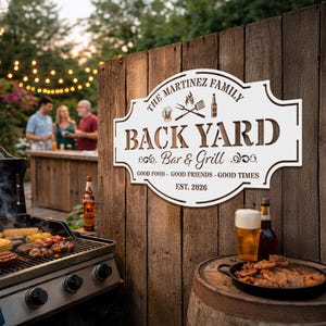 May include: A white metal sign with the text "THE MARTINEZ FAMILY BACK YARD Bar & Grill" is mounted on a wooden fence. The sign also includes the text "GOOD FOOD - GOOD FRIENDS - GOOD TIMES EST. 2026". A grill and food are in the foreground.