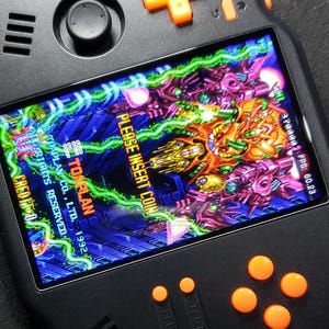 Puede incluir: Consola de juegos portátil negra con botones y cruceta naranja. La pantalla muestra un colorido juego de arcade con el texto "PLEASE INSERT COIN". La consola tiene el texto "TOAPLAN CO., LTD. 1992" en la pantalla.