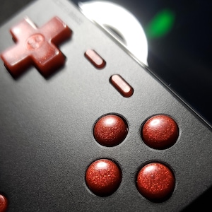 May include: A handheld gaming console with a matte black casing featuring a red glittery directional pad and four matching red glittery circular action buttons. The device includes two small oval buttons and a speaker grille on the front panel.