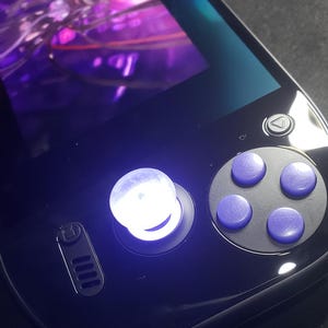May include: A black handheld gaming device with a glowing joystick and purple buttons. The screen displays a blurred, colorful graphic. The device has a sleek, modern design, with a directional pad and four action buttons.