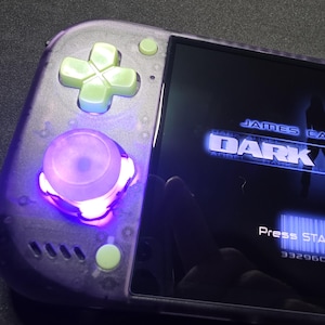 May include: A handheld gaming console with a translucent purple shell, light green directional pad and buttons, and a glowing purple analog stick. The screen displays the title James Cameron's Dark Angel with the text Press START button.