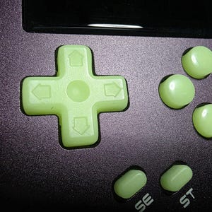 May include: Close-up of a handheld gaming device with a purple casing. The device features a light green directional pad and buttons, including a cross-shaped D-pad and four round buttons. The buttons are labeled "SE" and "ST".