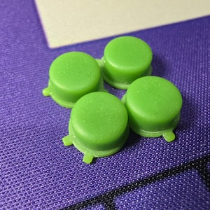 Puede incluir: Un conjunto de cuatro botones circulares pequeños de plástico color verde lima. Cada botón presenta una superficie superior lisa y plana con un borde ligeramente elevado y una pequeña lengüeta delgada que sobresale de la base.