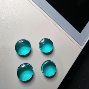 May include: Close-up of a white handheld gaming device with four teal-colored, circular buttons. The buttons are smooth and glossy, contrasting with the matte finish of the device. The screen is black.