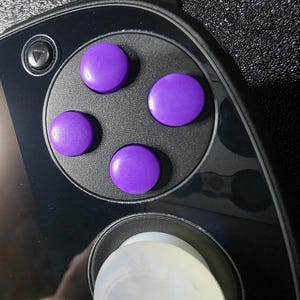 May include: Close-up of a black handheld gaming device with four purple circular buttons. The device has a sleek, modern design with a glossy finish. A directional button is visible on the upper left.
