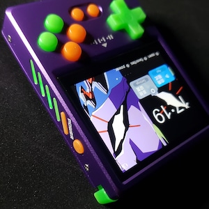 Puede incluir: Una consola de juegos portátil morada con botones verdes y naranjas. La pantalla muestra una imagen de anime colorida. El dispositivo tiene una cruceta y varios botones de control. La palabra "POWER" está impresa en el lateral.