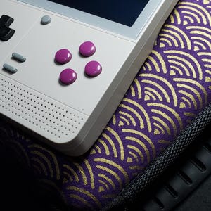 May include: A white handheld gaming console with purple buttons and a black screen rests on a purple and gold patterned case. The console has a directional pad, a small grey button, and a speaker.