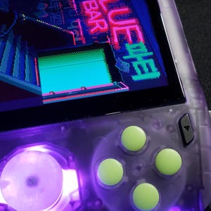 May include: A handheld games console with a screen showing a pixelated arcade game. The device has a translucent purple casing, green buttons, and a circular light. The screen displays the text "BLUE BAR".