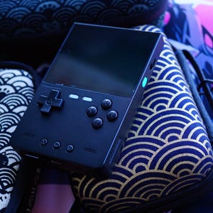May include: A black handheld gaming console with a D-pad and buttons, resting on a patterned surface. The console is surrounded by cases and accessories with wave and cat designs. The screen is dark, and a small light is visible.