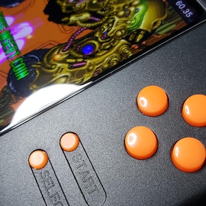 Könnte beinhalten: Ein Handheld-Spielgerät mit einem Bildschirm, der ein farbenfrohes Videospiel anzeigt. Das Gerät hat ein schwarzes Gehäuse mit orangefarbenen Knöpfen und den Aufschriften "SELECT" und "START" auf der Oberfläche.