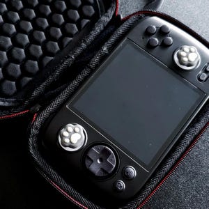 Op de afbeelding: Een zwarte handheld gameconsole met een groot scherm, knoppen en thumbstick-covers met pootafdrukken. De console zit in een zwarte etui met ritssluiting en een gestructureerd interieur.