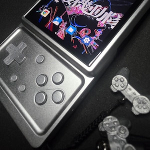 Pode incluir: Uma consola de jogos portátil prateada com um ecrã digital que apresenta um gráfico colorido de estilo anime e ícones de aplicações. O dispositivo possui um pad direcional e quatro botões circulares. Dois pequenos pendentes prateados em forma de comando estão anexados.