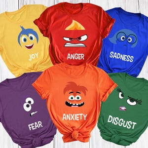 May include: A collection of six colorful t-shirts, each featuring a different character face and emotion from the movie "Inside Out." The shirts are yellow, red, blue, purple, orange, and green, with the words "Joy," "Anger," "Sadness," "Fear," "Anxiety," and "Disgust."