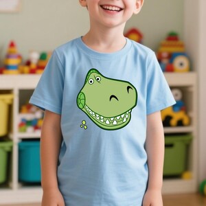 Peut inclure: Un t-shirt bleu clair avec un motif de t&ecirc;te de dinosaure verte de style dessin anim&eacute;. Le dinosaure a des dents blanches, des yeux noirs et un sourire courb&eacute;. Le t-shirt est en mati&egrave;re douce et convient aux enfants.