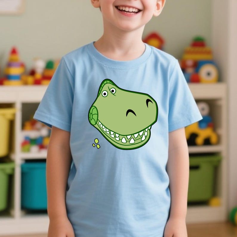 Peut inclure: Un t-shirt bleu clair avec un motif de t&ecirc;te de dinosaure verte de style dessin anim&eacute;. Le dinosaure a des dents blanches, des yeux noirs et un sourire courb&eacute;. Le t-shirt est en mati&egrave;re douce et convient aux enfants.