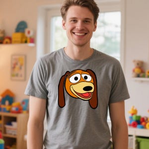 Peut inclure: Un t-shirt gris avec une image de dessin anim&eacute; de Slinky Dog de Toy Story. Le graphisme montre la t&ecirc;te du chien avec des oreilles marron, un museau jaune et une langue rouge. Le t-shirt est en mati&egrave;re souple.
