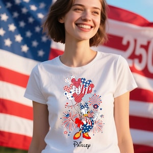 May include: White t-shirt featuring a patriotic design with Mickey Mouse-shaped balloons in red, white, and blue. The design includes fireworks, stars, and the text "Paisley". The shirt is set against a backdrop of the American flag.