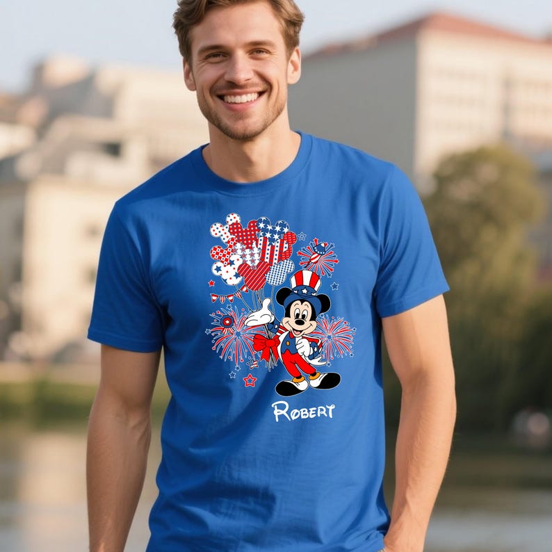 May include: A blue t-shirt featuring a cartoon Mickey Mouse in patriotic attire, holding balloons and fireworks. The design includes red, white, and blue elements, with the name "Robert" printed below the character.