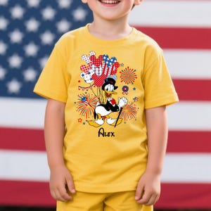 May include: Yellow t-shirt featuring a cartoon Donald Duck holding balloons and a cane, with fireworks and stars. The shirt has the name "Alex" printed below the character. The background is an American flag.