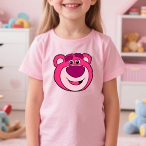 Peut inclure: Un t-shirt rose clair avec un visage d'ours de dessin anim&eacute; dans des tons de rose et de magenta. L'ours a un grand sourire et est centr&eacute; sur le t-shirt. Le t-shirt est port&eacute; par un jeune enfant.