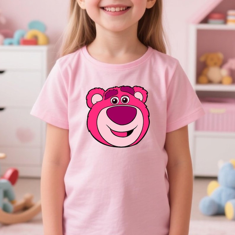 Peut inclure: Un t-shirt rose clair avec un visage d'ours de dessin anim&eacute; dans des tons de rose et de magenta. L'ours a un grand sourire et est centr&eacute; sur le t-shirt. Le t-shirt est port&eacute; par un jeune enfant.