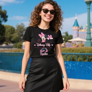 K&ouml;nnte beinhalten: Schwarzes T-Shirt mit einem Minnie Mouse-Motiv und dem Text "Disney Trip" und "Fapian" in Pink. Das Design umfasst Minnie Mouse, eine Schleife und Sonnenbrillen. Das T-Shirt hat einen Rundhalsausschnitt.