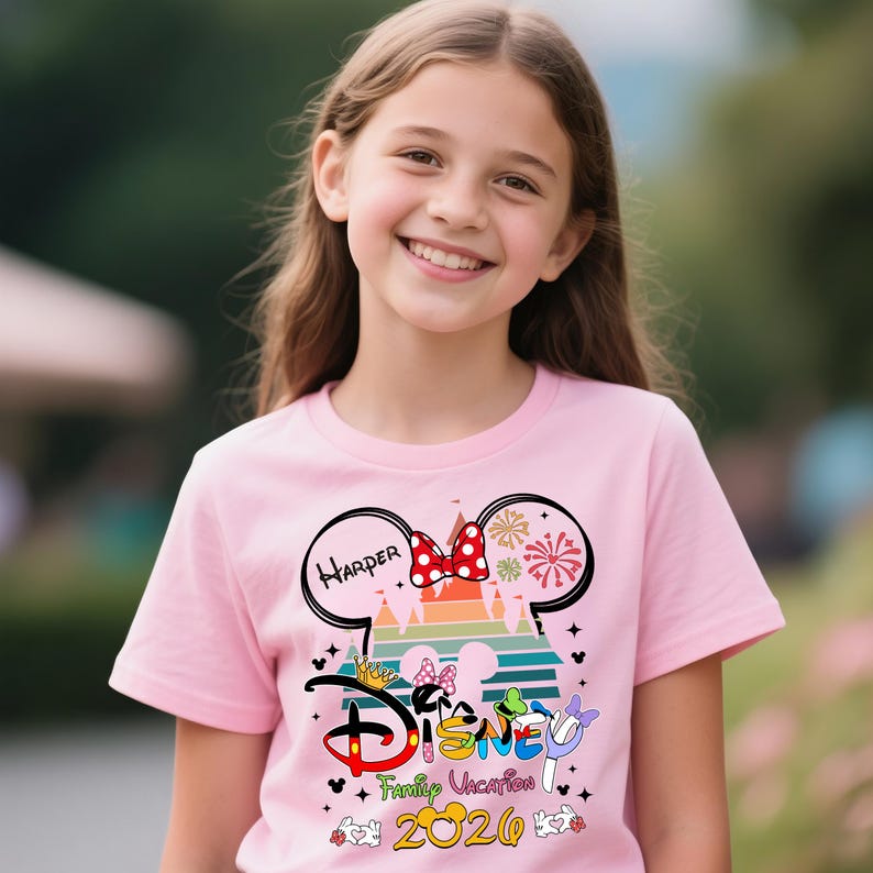 Pu&ograve; includere: T-shirt rosa con grafica a tema Disney. Il design include orecchie di Topolino, un fiocco rosso, fuochi d'artificio e la parola "Disney" in un font colorato. &Egrave; visibile anche il testo "Family Vacation 2020".
