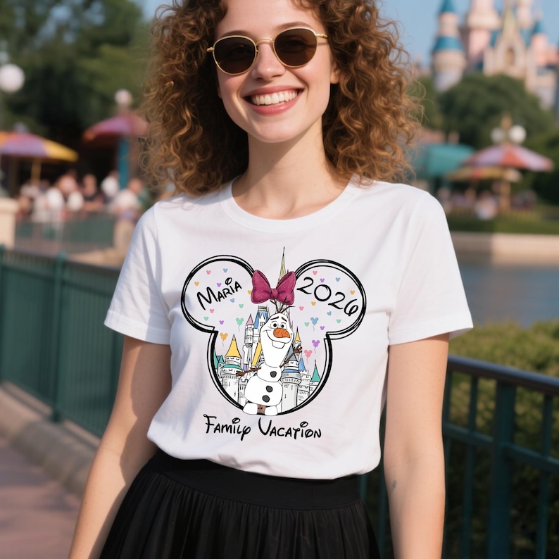 Puede incluir: Camiseta blanca con un dise&ntilde;o en forma de Mickey Mouse. El dise&ntilde;o incluye el nombre "Maria", el a&ntilde;o "2026", un dibujo animado de Olaf, un castillo y las palabras "Family Vacation". Un recuerdo de vacaciones familiares.