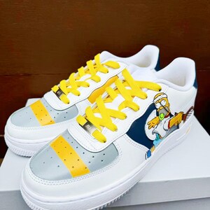 以下が含まれることがあります： 黄色い靴ひもが付いた白いスニーカーで、漫画のキャラクターのデザインが施されています。キャラクターは青いシャツと黄色のショートパンツを着て、テニスラケットを持っています。スニーカーには、側面にグレーと黄色のストライプがあります。