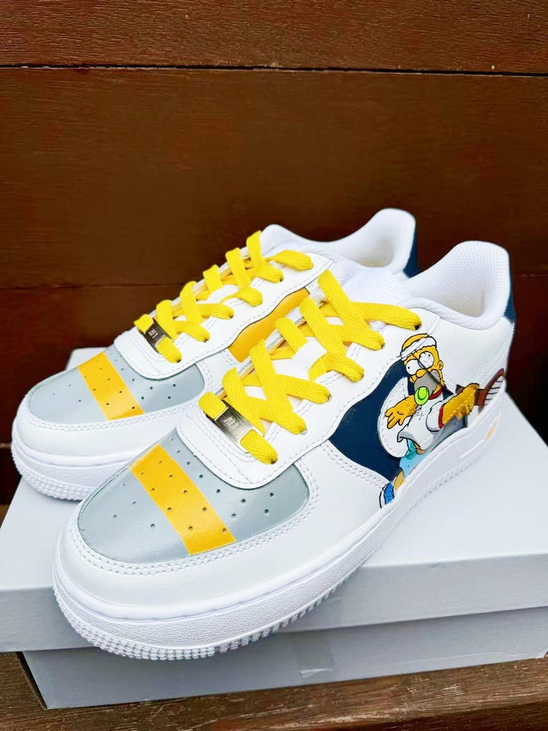 以下が含まれることがあります： 黄色い靴ひもが付いた白いスニーカーで、漫画のキャラクターのデザインが施されています。キャラクターは青いシャツと黄色のショートパンツを着て、テニスラケットを持っています。スニーカーには、側面にグレーと黄色のストライプがあります。