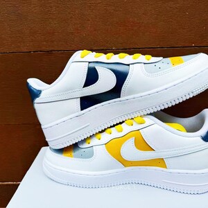 以下が含まれることがあります： 黄色と青のアクセントが付いた白いスニーカーのペア。スニーカーは白い革のアッパーに黄色のスウッシュと青いアクセントがヒールに付いています。スニーカーは白い表面の上にあります。