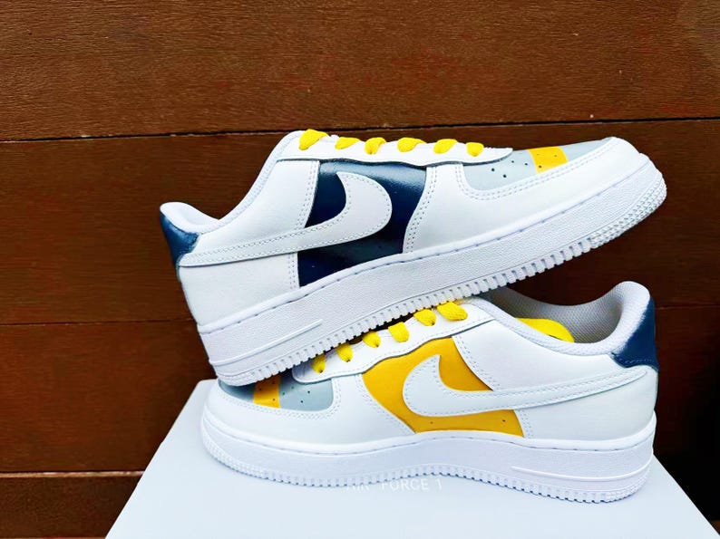 以下が含まれることがあります： 黄色と青のアクセントが付いた白いスニーカーのペア。スニーカーは白い革のアッパーに黄色のスウッシュと青いアクセントがヒールに付いています。スニーカーは白い表面の上にあります。