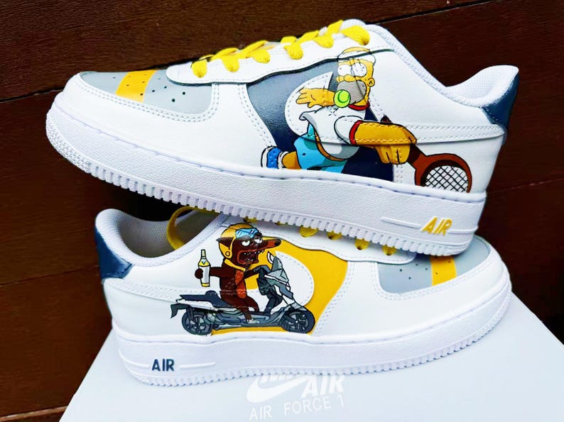 以下が含まれることがあります： 黄色、青、グレーのアクセントが付いたカスタムペイントされた白いナイキエアフォース1スニーカー。片方の靴にはテニスをしている漫画のキャラクターが描かれ、もう片方の靴にはスクーターに乗っている漫画のキャラクターが描かれています。