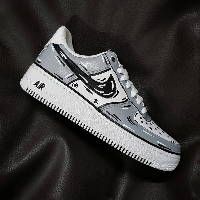 Może przedstawiać: Szare i czarne, spersonalizowane buty Nike Air Force 1 z wzorem w stylu kresk&oacute;wkowym. Buty mają czarne logo Swoosh i napis "AIR" na boku.