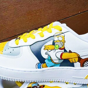以下が含まれることがあります： 黄色い靴ひもが付いた白いスニーカーで、テニスをしているカートゥーンのホーマー・シンプソンのイラストが描かれています。スニーカーには青と灰色のスウッシュロゴと、側面に「AIR」の文字が印刷されています。