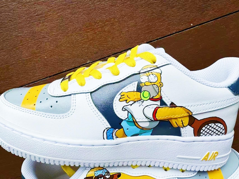 以下が含まれることがあります： 黄色い靴ひもが付いた白いスニーカーで、テニスをしているカートゥーンのホーマー・シンプソンのイラストが描かれています。スニーカーには青と灰色のスウッシュロゴと、側面に「AIR」の文字が印刷されています。