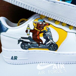 以下が含まれることがあります： 黄色と青の模様が施された白い革のスニーカー。漫画のキャラクターがスクーターに乗っているデザインです。スニーカーには側面に「AIR FORCE 1」の文字があります。