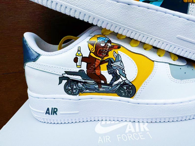 以下が含まれることがあります： 黄色と青の模様が施された白い革のスニーカー。漫画のキャラクターがスクーターに乗っているデザインです。スニーカーには側面に「AIR FORCE 1」の文字があります。