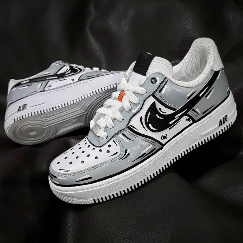 Może przedstawiać: Białe buty Nike Air Force 1, pomalowane na zam&oacute;wienie, z czarnymi i szarymi grafikami w stylu kresk&oacute;wkowym. Buty mają białą podeszwę środkową i czarną podeszwę zewnętrzną. Tekst "AIR" jest nadrukowany na boku but&oacute;w.