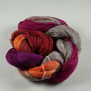 May include: A skein of colorful yarn in shades of purple, red, orange, and gray. The yarn is tightly wound and appears to be made of a soft, shiny material. The colors blend together, creating a visually appealing texture.