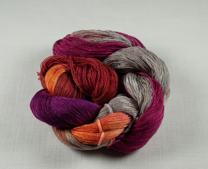 May include: A skein of colorful yarn in shades of purple, red, orange, and gray. The yarn is tightly wound and appears to be made of a soft, shiny material. The colors blend together, creating a visually appealing texture.