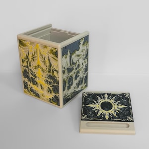 May include: A beige rectangular storage box featuring detailed illustrations of architectural structures in yellow, black, and white. The detachable lid displays a central sun-like emblem with radiating points, set against a dark, textured background.