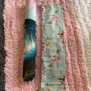 May include: A rolled-up yoga mat with a watercolor ocean design and a floral print carrying bag. The bag is light green with pink and yellow roses. The mat and bag are on a pink and white fluffy surface.