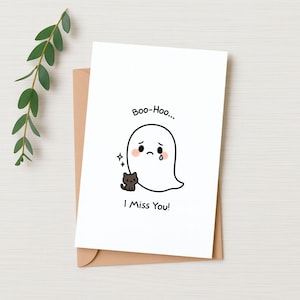 May include: A white greeting card with a sad ghost illustration, a small black cat, and the text "Boo-Hoo... I Miss You!". The card is on a brown envelope, with a sprig of green leaves in the background.