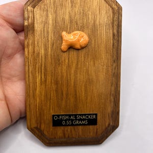 May include: A wooden plaque with a small, orange, fish-shaped object affixed to it. The plaque has a dark wood grain finish and a rectangular shape with beveled edges. A small black label reads "O-FISH-AL SNACKER 0.55 GRAMS".