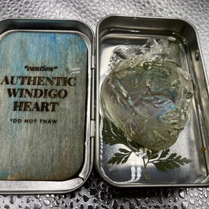 May include: An open metal tin containing a translucent, heart-shaped object and a wooden insert. The insert reads "*caution* AUTHENTIC WINDIGO HEART *DO NOT THAW" in dark brown text. The heart rests on green foliage.