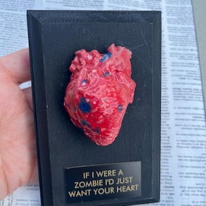Pode incluir: Escultura de um coração vermelho e azul montada numa placa de madeira preta. A placa tem uma placa de metal com o texto "IF I WERE A ZOMBIE I'D JUST WANT YOUR HEART".