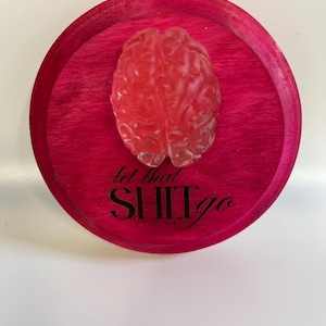 Puede incluir: Una placa redonda de madera rosa intenso con un objeto translúcido en forma de cerebro rojo en el centro. La frase "let that SHIT go" está impresa en escritura negra debajo del cerebro.
