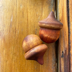 May include: Two wooden acorns with a brown and reddish-brown finish. The acorns are sitting on a wooden surface.